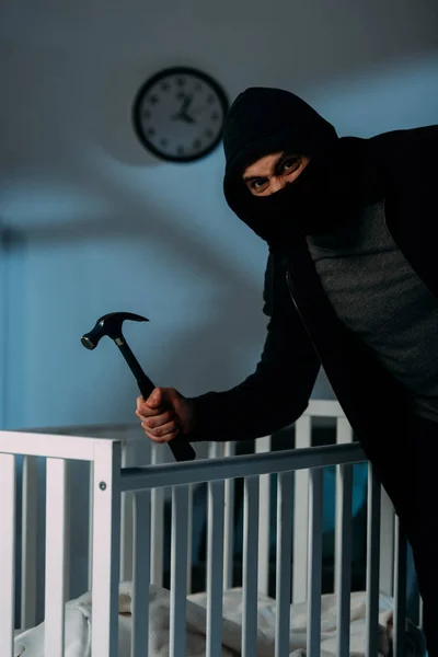 Angry criminal in mask standing near crib and holding hammer — Stock Photo