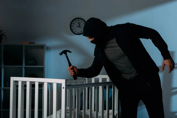 Преступник в маске держит молоток, стоя возле кроватки в темной комнате — стоковое фото