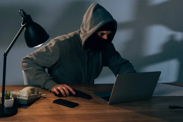 Criminal in mask with gun using laptop in dark room — Stock Photo