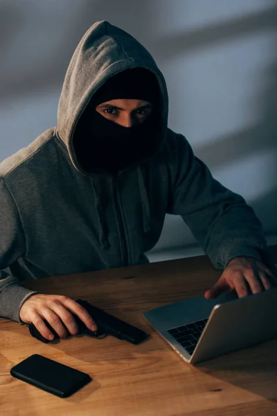 Verbrecher maskiert mit Waffe und Laptop im dunklen Raum — Stockfoto