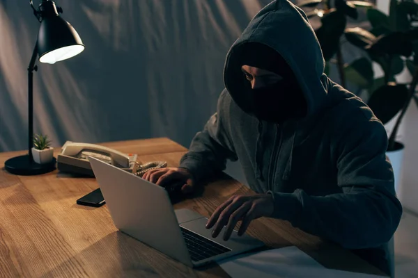 Verbrecher mit Maske und Kapuzenpulli tippt auf Laptop-Tastatur im dunklen Raum — Stockfoto