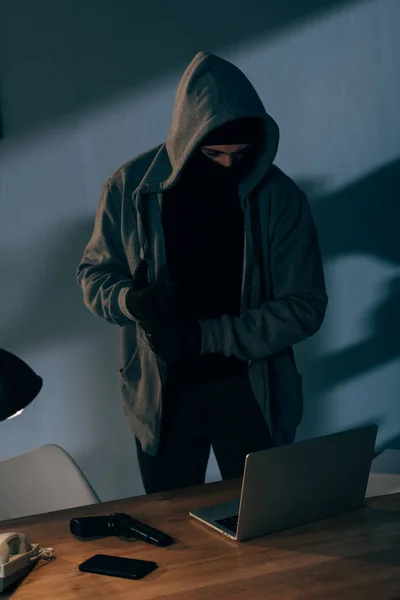 Criminal in mask and hoodie looking at laptop in dark room — Stock Photo