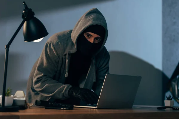 Cambrioleur masqué et gants tapant sur un clavier d'ordinateur portable dans une pièce sombre — Photo de stock