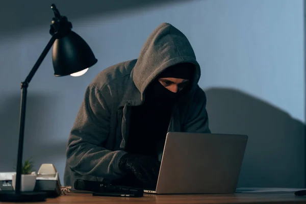 Criminal en máscara y sudadera con capucha usando portátil en habitación oscura - foto de stock