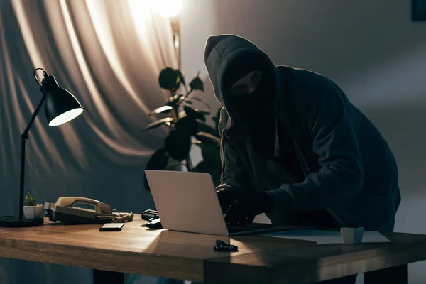 Verbrecher in Kapuzenpulli und Maske mit Laptop im dunklen Raum — Stockfoto