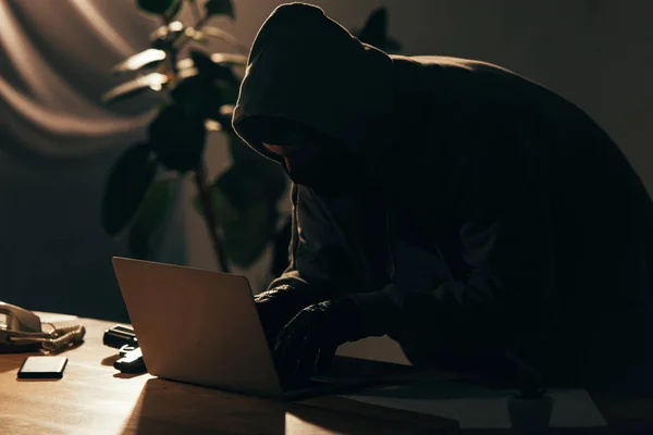 Criminal in hoodie and mask using laptop in dark room — Stock Photo