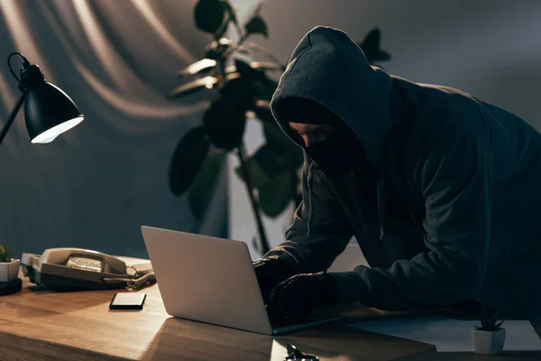 Criminal en sudadera con capucha y guantes escribiendo en el teclado del ordenador portátil en habitación oscura - foto de stock