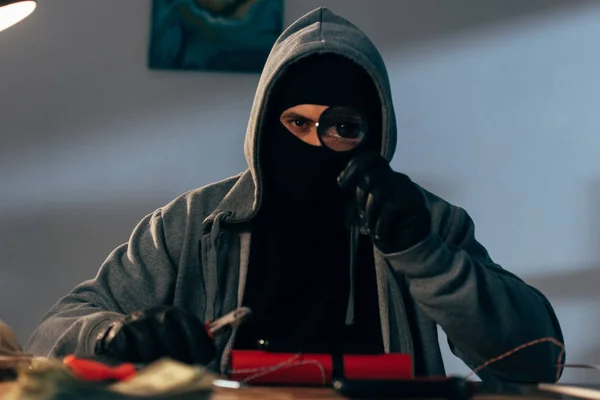 Terrorist in mask holding magnifier while making bomb in room — Stock Photo