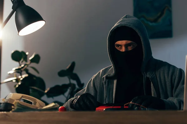Pensive терорист в масці і шкіряні рукавички сидять за столом з телефоном — стокове фото
