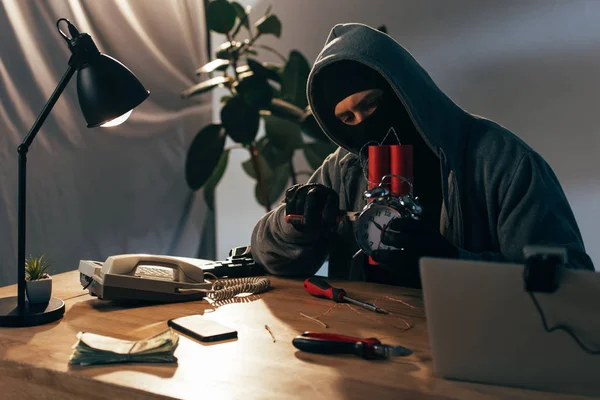 Criminal in mask sitting at table with laptop and making bomb — Stock Photo