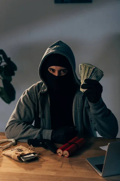 Terrorist in mask and gloves sitting at table with weapon and holding dollar banknotes — Stock Photo