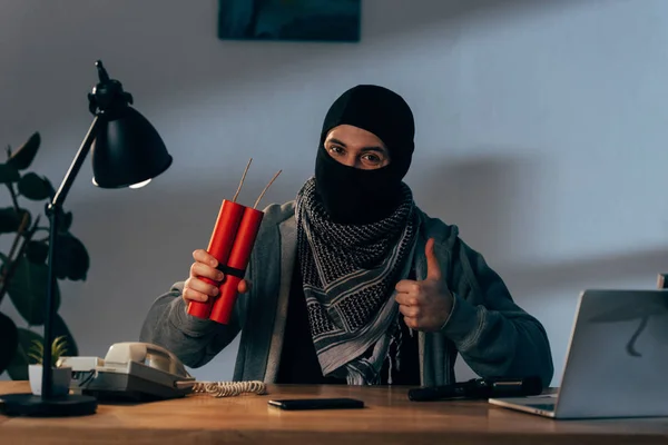 Terrorist in mask holding dynamite and showing thumb up in room — Stock Photo