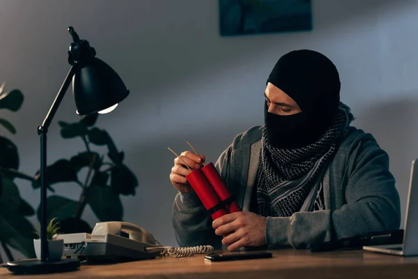 Terrorist in mask with dynamite sitting at table in room — Stock Photo