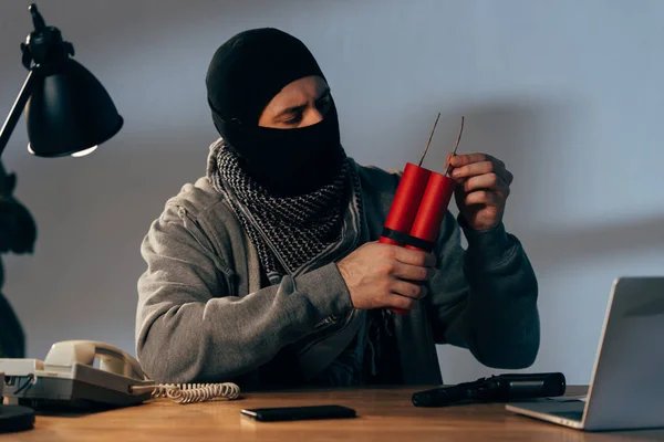 Terrorist mit Maske hält Dynamit in der Hand, während er im dunklen Raum am Tisch sitzt — Stockfoto