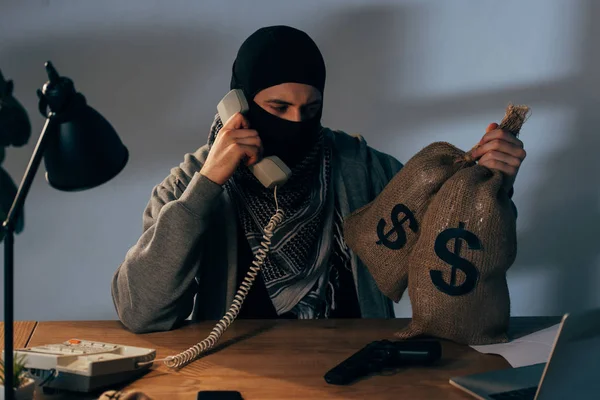 Terrorista en máscara sosteniendo bolsas con dinero y hablando por teléfono en la habitación - foto de stock
