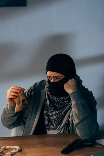 Triste terrorista in maschera guardando piccolo sacchetto di denaro in camera — Foto stock