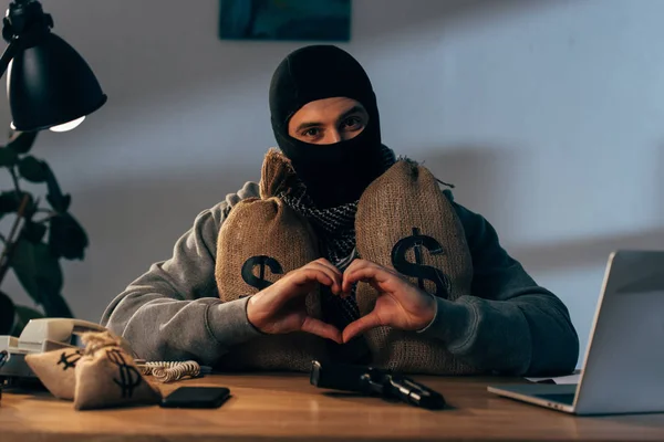 Terrorista en máscara con bolsas de dinero mostrando el signo del corazón - foto de stock