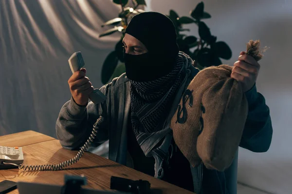 Angry terrorist in mask holding money bags and looking at handset — Stock Photo