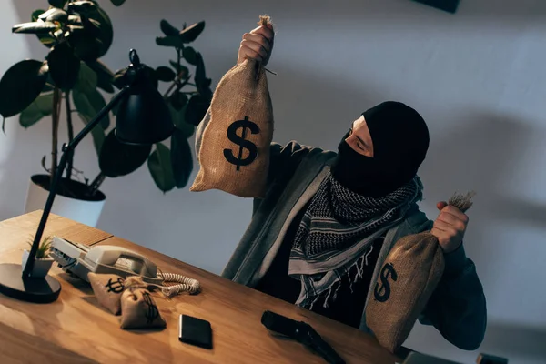 Terrorist in mask sitting at table and holding bags with dollars — Stock Photo