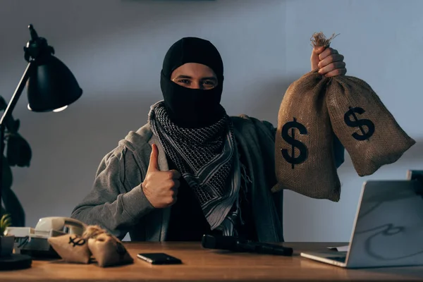 Terrorista en máscara sosteniendo bolsas con dólares y mostrando el pulgar hacia arriba - foto de stock