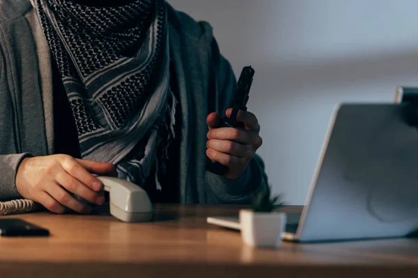 Partial view of terrorist holding handset and gun at table — Stock Photo