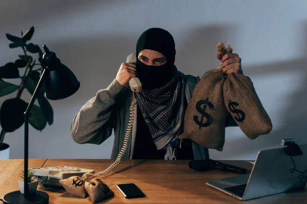 Злий терорист тримає сумки з доларовими знаками і розмовляє по телефону — стокове фото