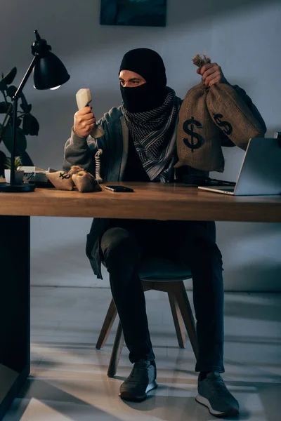 Terrorista agresivo con bolsas de dinero sentado en la habitación y sosteniendo el teléfono - foto de stock