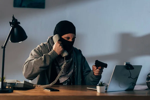 Angry terrorist in mask talking on telephone and showing gun at webcam — Stock Photo