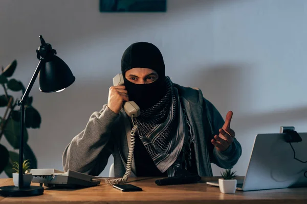 Terrorista enojado con máscara negra hablando por teléfono en la habitación - foto de stock