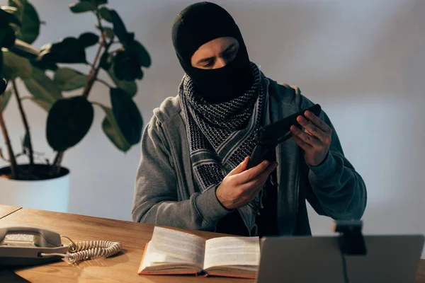 Nachdenklicher Terrorist mit schwarzer Maske blickt auf Waffe im Zimmer — Stockfoto