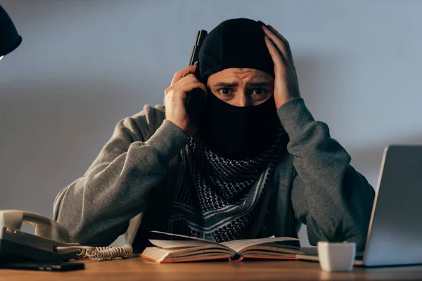 Worried terrorist in mask with book and gun touching head — Stock Photo