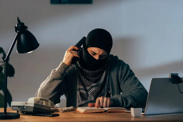 Terroriste concentré masqué avec livre de lecture d'armes dans la chambre — Photo de stock