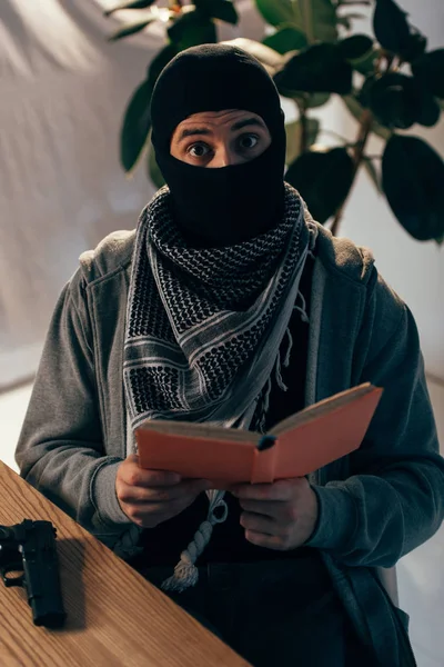 Schockierter Terrorist mit schwarzer Maske liest Buch am Tisch — Stockfoto