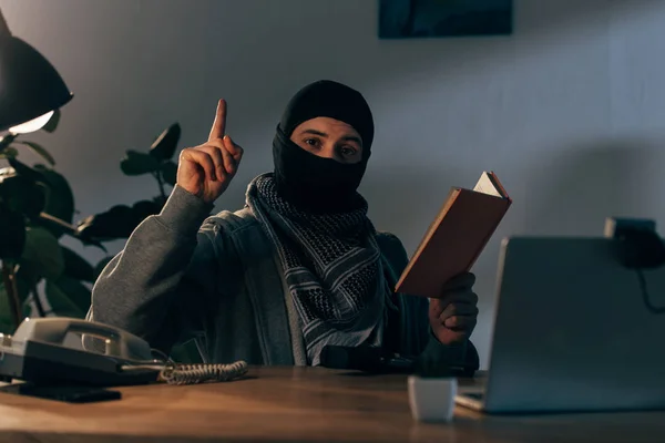 Terrorist in mask reading book and pointing with finger — Stock Photo