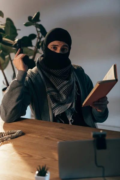 Terrorista de máscara preta segurando arma e lendo livro — Fotografia de Stock