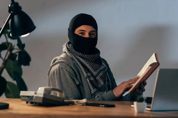 Terrorista con máscara negra sentado a la mesa y leyendo libro - foto de stock