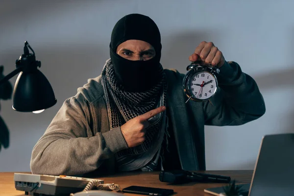 Angry terrorist in black mask pointing with finger at alarm clock — Stock Photo