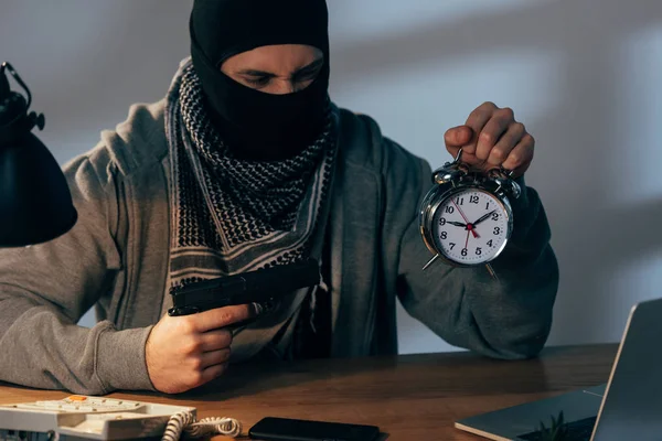 Terrorista enojado con máscara sosteniendo pistola y mirando el despertador - foto de stock