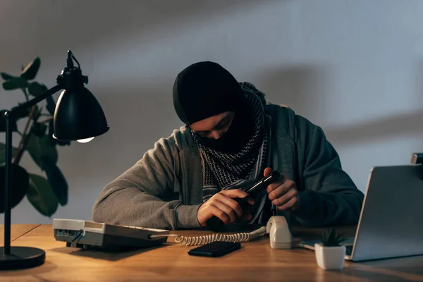 Terrorist mit schwarzer Maske sitzt am Tisch und hält Waffe — Stockfoto