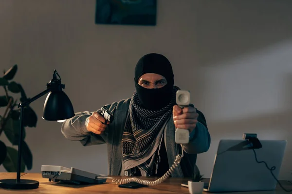 Агрессивный террорист в маске держит телефон и целится в пистолет — стоковое фото