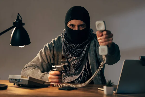 Angry terrorist in black mask holding gun and showing handset at camera — Stock Photo