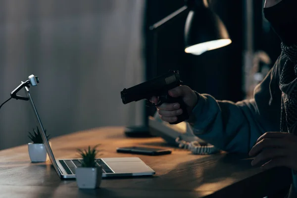 Ausgeschnittene Ansicht eines maskierten Verbrechers, der mit einer Waffe auf den Laptop zielt — Stockfoto