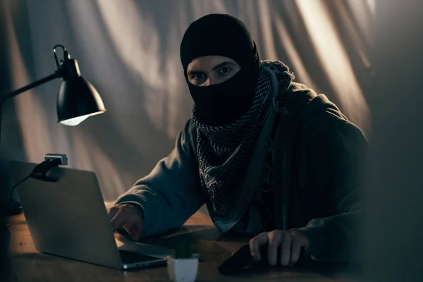 Terrorist in mask with gun using laptop in dark room — Stock Photo