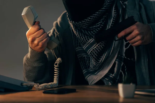 Cropped view of terrorist holding handset and gun at table — Stock Photo