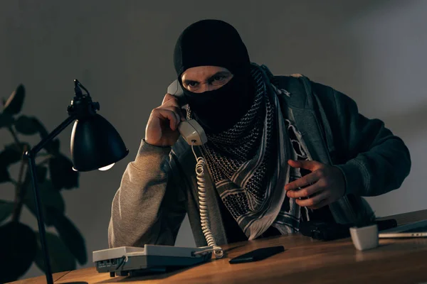 Terrorista agresivo con máscara negra sentado a la mesa y hablando por teléfono - foto de stock