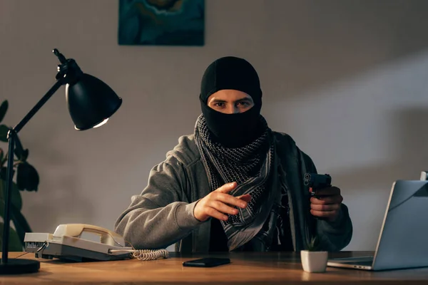 Террорист в маске сидит за столом с лампой и держит пистолет — стоковое фото