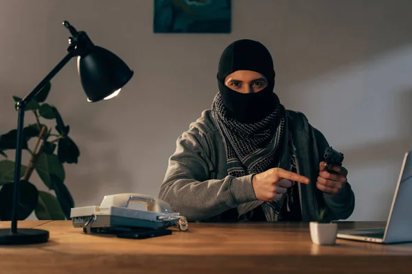 Terrorista de máscara preta sentado à mesa e apontando com o dedo para a arma — Fotografia de Stock