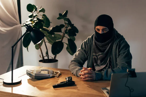 Criminal pensativo con máscara negra sentado en la mesa con los dedos entrelazados - foto de stock