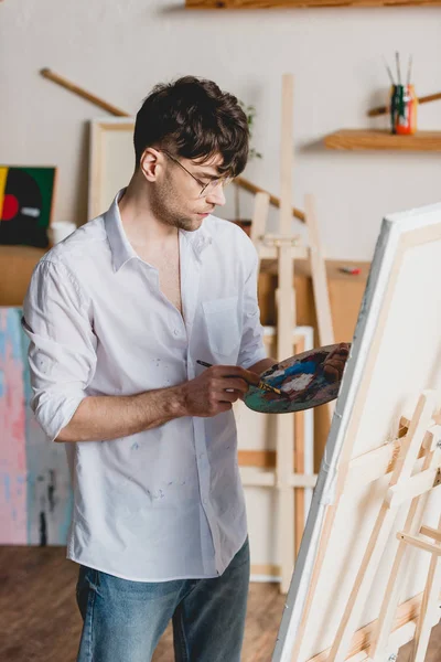 Красивый художник смешивает краски на палитре, стоя возле мольберта с холстом — стоковое фото