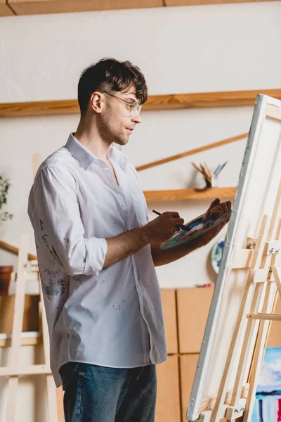 Good-looking artist mixing paints on palette while standing near easel with canvas — Stock Photo